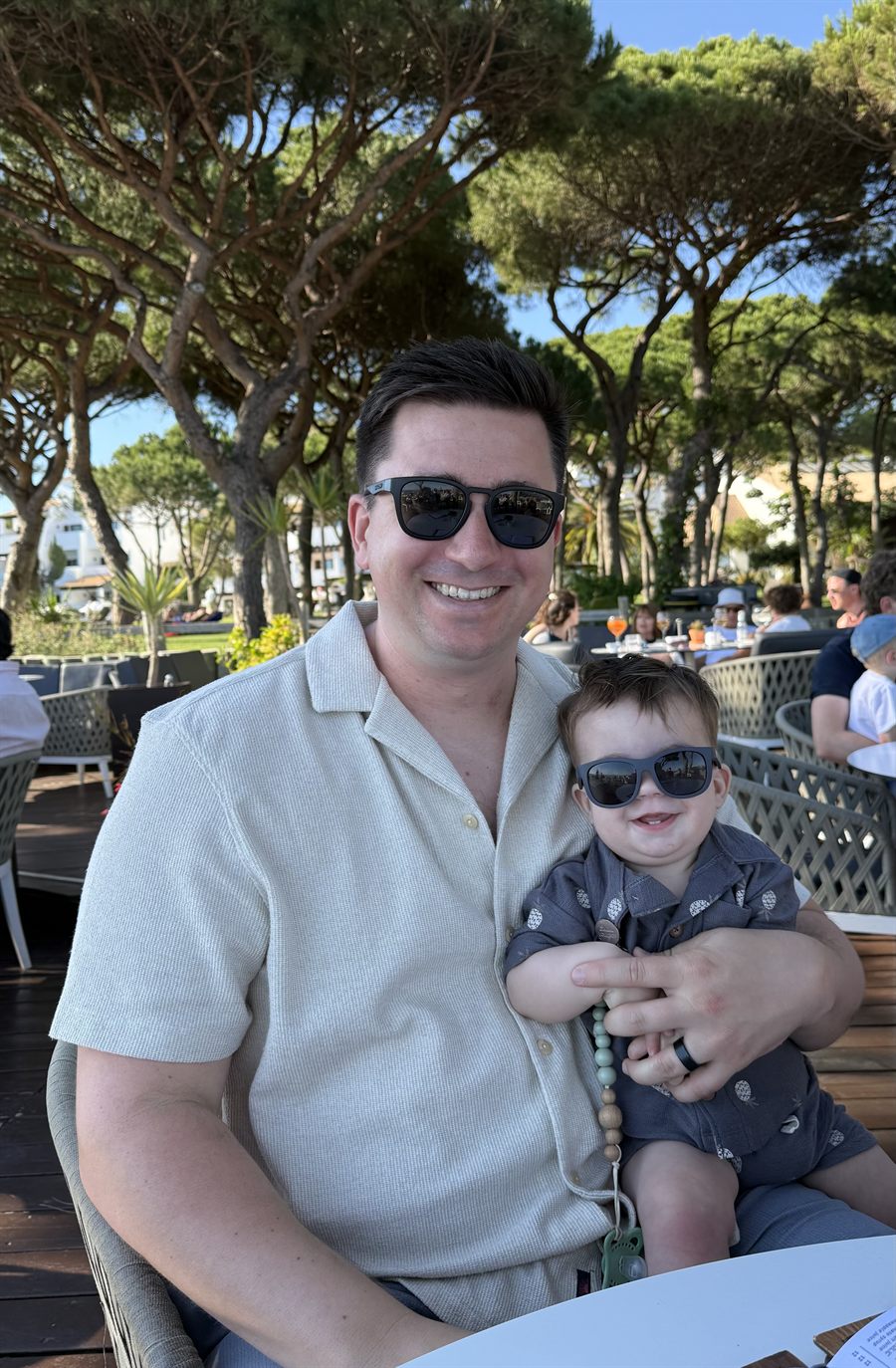 Pete on a shaded cafe terrace under tall pines, holding a smiling toddler; both are wearing sunglasses.